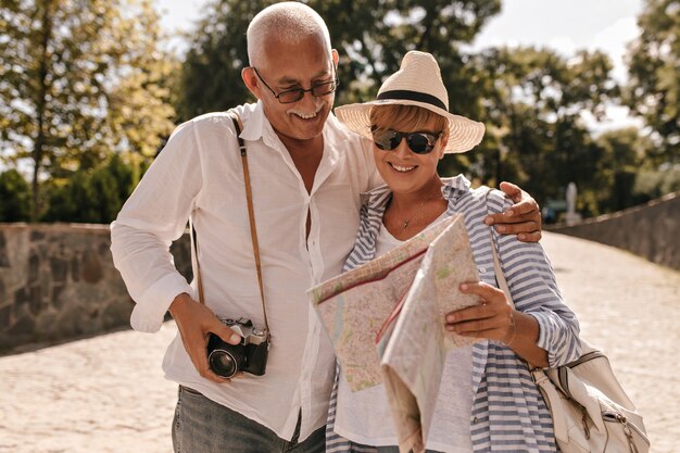 Homme gai aux cheveux gris en chemise légère et jeans avec caméra en souriant et en regardant la carte avec une dame blonde au chapeau et une tenue bleue dans le parc.