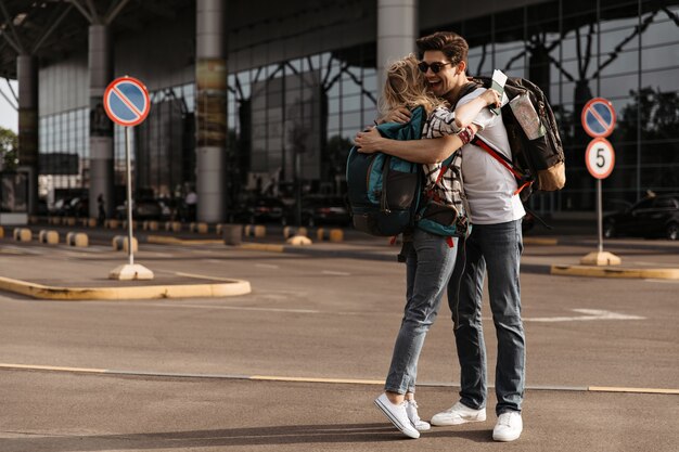 Homme gai dans des lunettes de soleil, tee-shirt blanc, câlins de jeans avec une femme blonde en chemise à carreaux