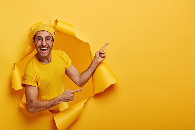 Un homme gai fait une belle offre, annonce un nouveau produit en vente, se tient dans un trou de papier déchiré, a une expression positive