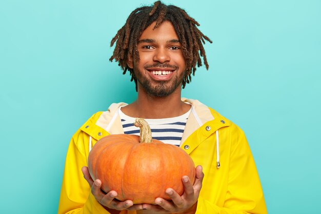 Homme gai en imperméable jaune se vante de sa récolte d'automne, tient la citrouille, sourit agréablement, pose sur le mur bleu