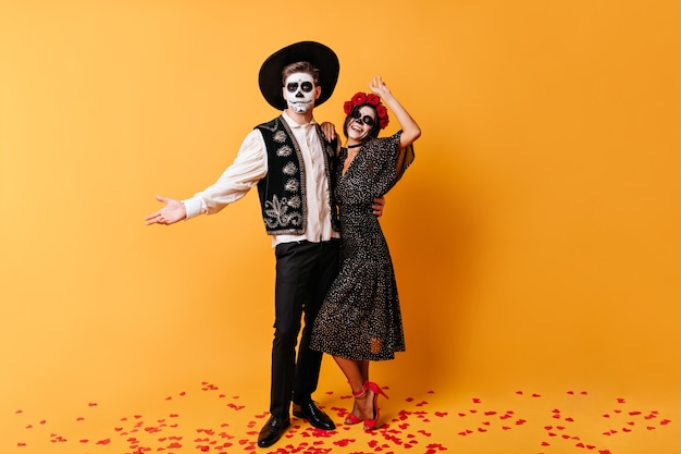 Homme gai en sombrero et sa femme élégante avec du maquillage d'Halloween posant joyeusement, serrant contre le mur orange.