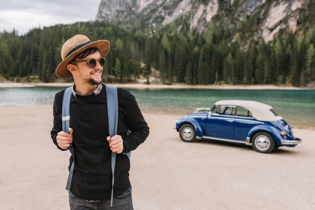 Homme gai tenant son sac à dos et posant avec le sourire debout devant le magnifique lac sur la montagne