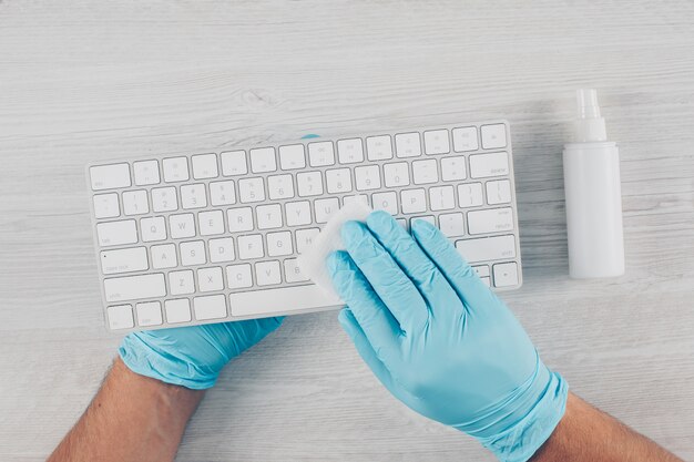 Un homme en gants désinfectant un clavier en fond de bois clair avec un désinfectant.