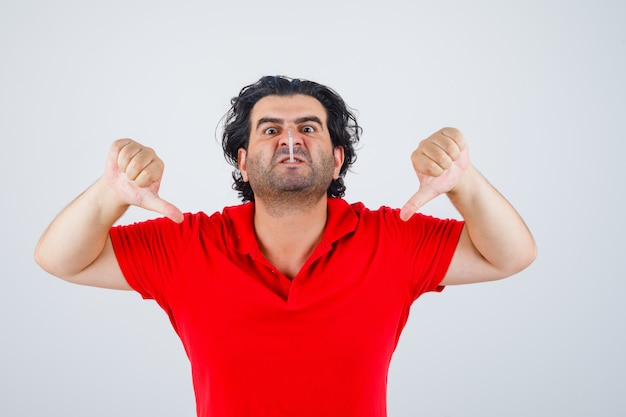 Homme gardant la cigarette dans la bouche, montrant le double pouce vers le bas en t-shirt rouge et l'air furieux.