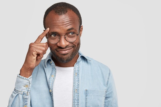 L'homme garde l'index sur les tempes, essaie de se concentrer sur quelque chose, habillé en chemise en jean, isolé sur un mur blanc