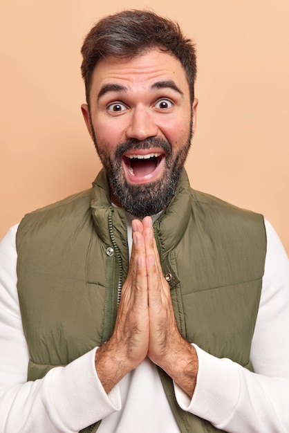 Photo gratuite l'homme garde les paumes pressées l'une contre l'autre croit en quelque chose de bon espère que quelque chose de bien s'est produit vêtu d'un pull blanc et d'un gilet isolé sur beige