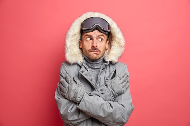 Un homme glacé perplexe croise les mains et essaie de se réchauffer tremble de froid par temps neigeux à basse température porte des gants de veste d'hiver chauds et des lunettes de ski tremblant de gel par temps glacial