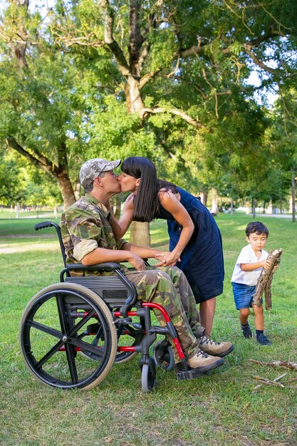 Homme handicapé en uniforme militaire embrassant sa femme pendant que leur petit péché transportait du bois de chauffage pour feu de camp dans le parc. Ancien combattant handicapé ou concept extérieur familial