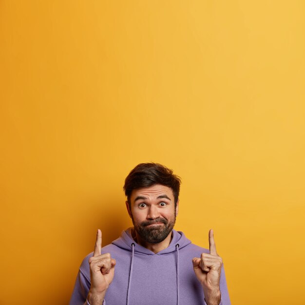 Homme hésitant perplexe avec barbe, presse les lèvres, porte un sweat à capuche violet, pointe l'index au-dessus, hésite quoi acheter, a une expression désemparée, isolé sur un mur jaune, espace vide vers le haut