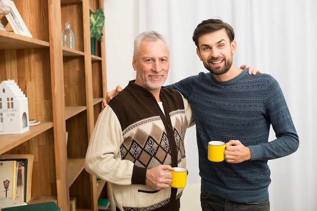 Photo gratuite homme heureux âgé embrassant avec jeune homme avec des tasses dans la chambre