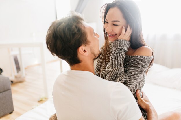 Homme heureux aux cheveux courts embrassant la jeune femme en riant dans des vêtements doux gris