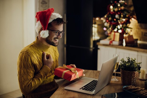 Homme heureux avec boîte-cadeau faisant un appel vidéo sur un ordinateur portable la veille de Noël à la maison