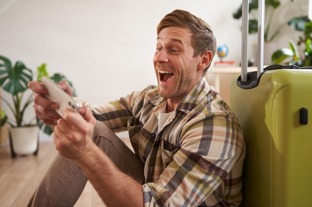 Un homme heureux jouant sur son téléphone portable, riant et souriant, assis avec une valise pour partir en vacances.