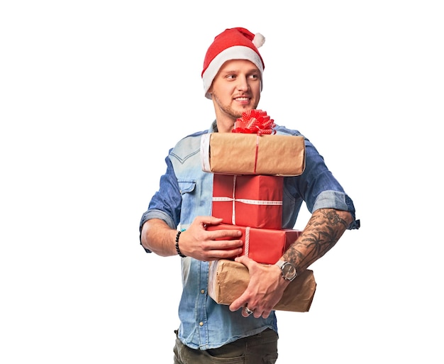 Un homme heureux vêtu d'une chemise en jean et d'un bonnet de Noel tenant des boîtes-cadeaux. Isolé sur fond blanc.