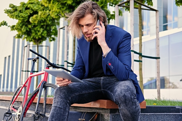 Photo gratuite un homme hipster aux longs cheveux blonds parle par téléphone intelligent et tient une tablette pc avec un vélo à vitesse unique en arrière-plan.