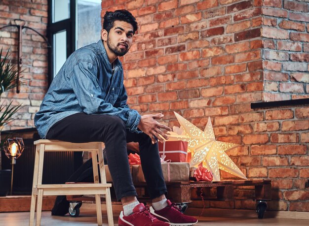 Homme hipster barbu indien avec des tatouages sur le bras dans une chambre loft avec décoration de Noël.