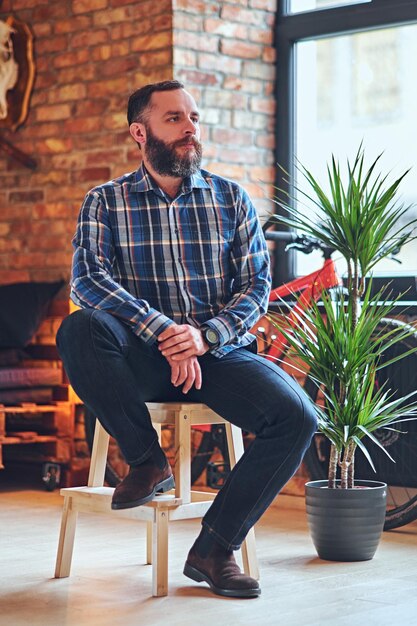 Un homme hipster barbu vêtu d'une chemise polaire est assis sur une chaise en bois, près de la fenêtre dans une pièce à l'intérieur loft