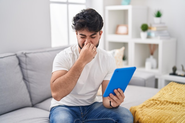 Photo gratuite homme hispanique avec barbe utilisant un pavé tactile assis sur le canapé sentant quelque chose de puant et dégoûtant odeur intolérable retenant son souffle avec les doigts sur le nez mauvaise odeur