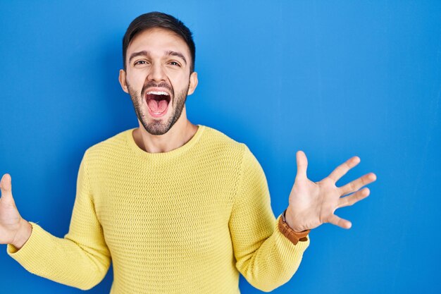 Homme hispanique debout sur fond bleu célébrant fou et étonné du succès avec les bras levés et les yeux ouverts criant le concept de gagnant excité