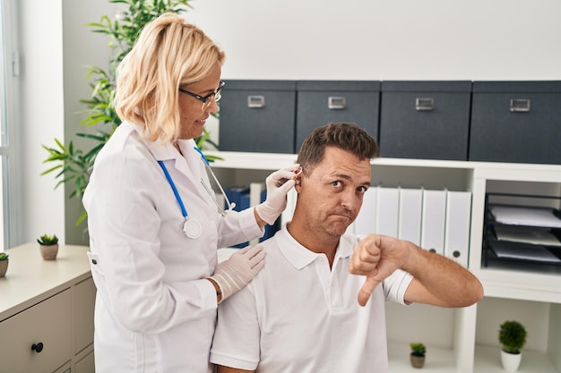 Photo gratuite homme hispanique recevant une aide auditive médicale chez le médecin avec un visage en colère, signe négatif montrant l'aversion avec les pouces vers le bas, concept de rejet