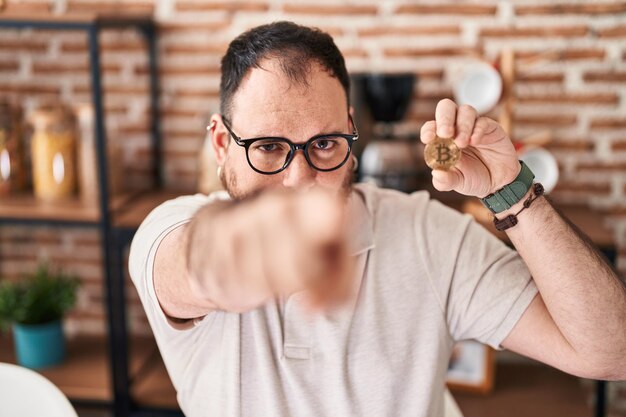 Homme hispanique de taille plus avec barbe tenant un bitcoin de monnaie virtuelle pointant du doigt vers l'appareil photo et vers vous un geste confiant à la recherche de sérieux