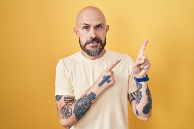 Photo gratuite homme hispanique avec des tatouages debout sur fond jaune pointant de côté inquiet et nerveux avec les deux mains concernées et expression surprise