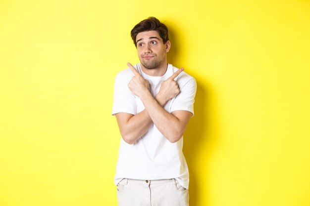 Homme indécis haussant les épaules, pointant du doigt sur le côté, soucieux de choisir, debout sur fond jaune.