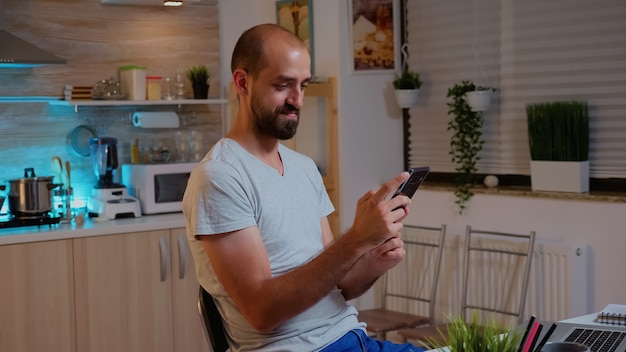 Homme indépendant utilisant le téléphone et souriant tout en faisant des heures supplémentaires à domicile. Employé concentré occupé utilisant un réseau de technologie moderne sans fil tard dans la nuit, lecture, écriture, recherche