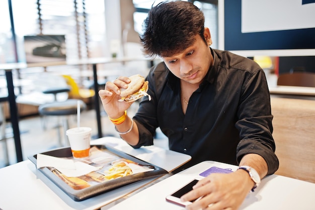 Un homme indien élégant assis dans un café de restauration rapide et mangeant un hamburger lit les nouvelles du matin sur son téléphone portable et a des yeux surpris