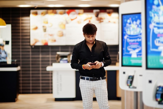 Photo gratuite homme indien élégant posé dans un café de restauration rapide près d'un kiosque d'auto-paiement avec téléphone portable à portée de main