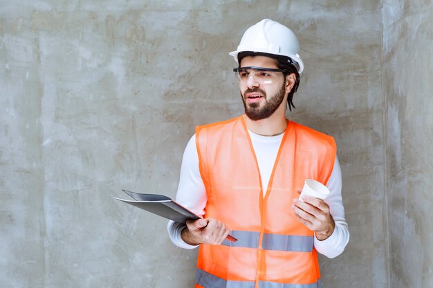 Homme ingénieur en casque blanc et lunettes de protection tenant un dossier noir et une tasse de boisson.