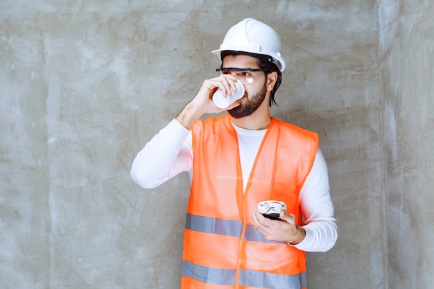 Homme ingénieur en casque blanc et lunettes de protection tenant un réveil et buvant une tasse de thé.