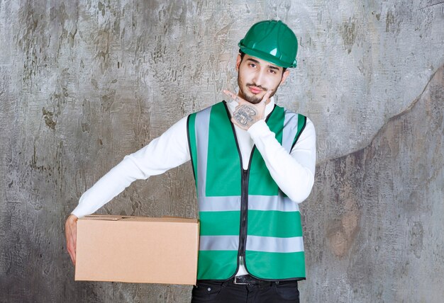 Homme ingénieur en uniforme jaune et casque tenant un colis en carton et a l'air réfléchi.