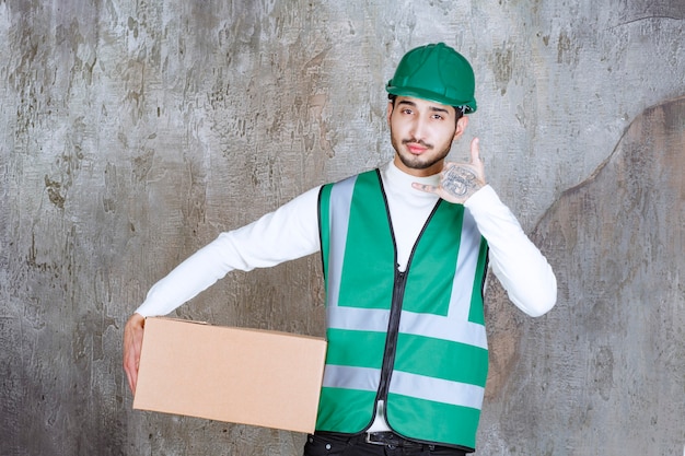 Homme ingénieur en uniforme jaune et casque tenant un colis en carton et demandant un appel.