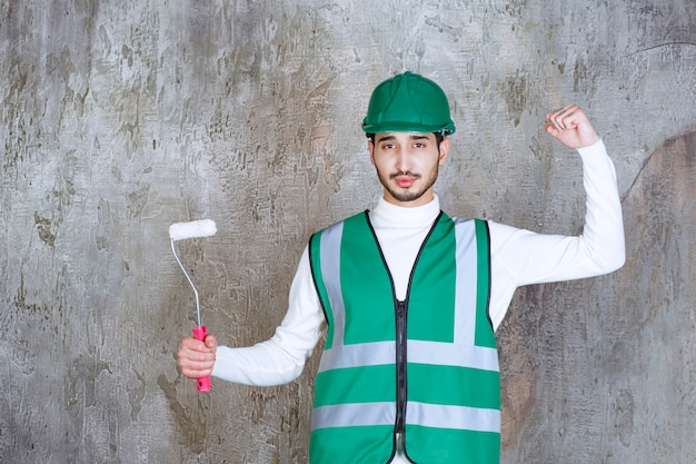 Homme ingénieur en uniforme jaune et casque tenant un rouleau de finition pour la peinture murale et montrant un signe positif de la main.