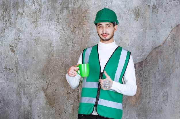 Homme ingénieur en uniforme jaune et casque tenant une tasse de café vert et appréciant le produit.