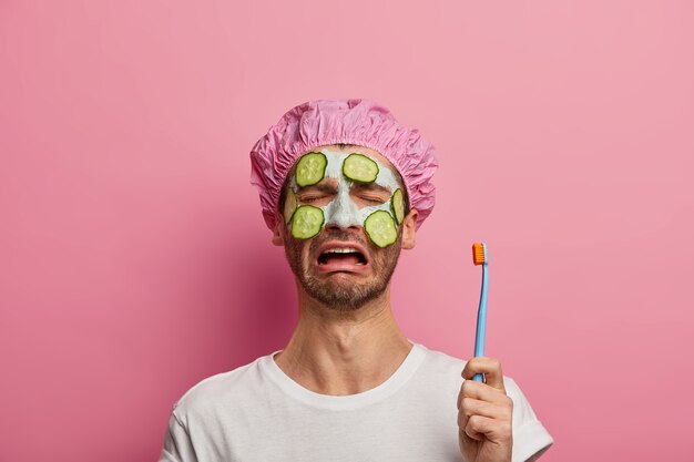 Un homme insatisfait qui pleure tient une brosse à dents, applique un masque d'argile pour le visage avec des concombres, fatigué des traitements de beauté