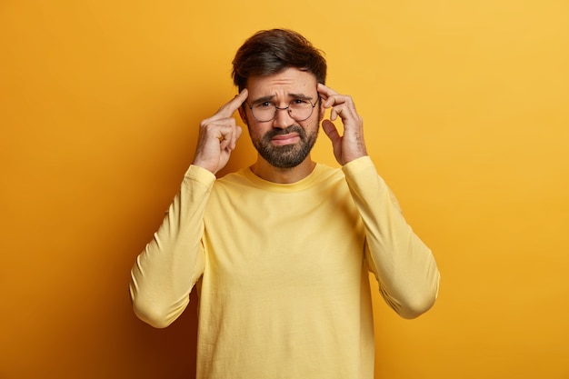 Un homme insatisfait réfléchit intensément, touche les tempes avec l'index, sourit narquoisement, souffre de maux de tête insupportables, porte un pull décontracté, pose à l'intérieur ressent de la pression et de la détresse, isolé sur jaune