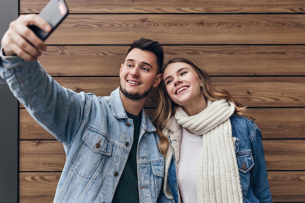 Homme inspiré avec barbe faisant selfie avec sa petite amie. Superbe jeune femme avec un foulard noir posant sur un mur en bois.