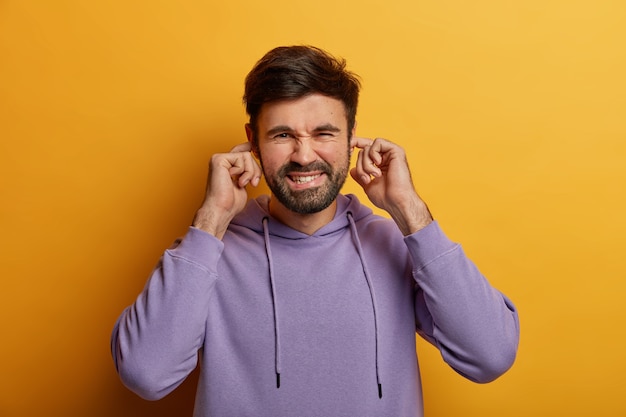 Un homme irrité et mécontent se bouche les oreilles, ne supporte pas le bruit ou le son, ignore les conflits, porte un sweat à capuche violet, isolé sur un mur jaune. Concept de langage corporel. Le jeune ne veut pas entendre de musique