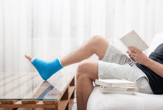 Un homme avec une jambe cassée dans un plâtre lit des livres sur un fond clair de l'intérieur de la pièce.