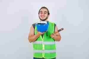 Photo gratuite homme jeune constructeur montrant un marteau et un casque en uniforme de vêtements de travail et à la joyeuse vue de face.