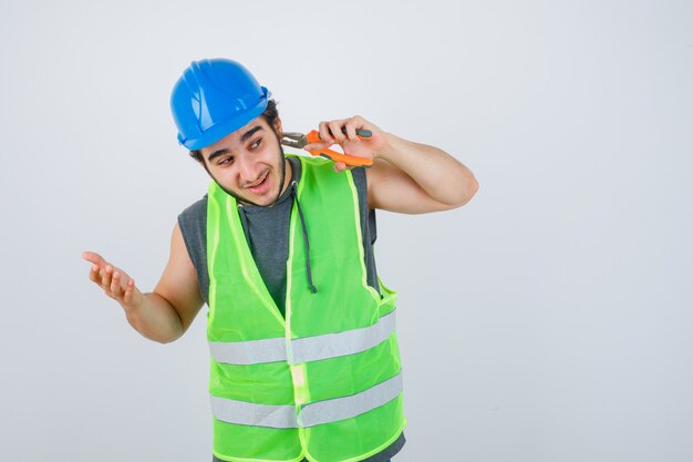 Homme Jeune Constructeur Pinçant L'oreille Avec Des Pinces En Uniforme De Vêtements De Travail Et à La Vue Amusée, De Face.