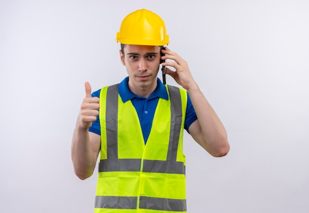 Photo gratuite homme jeune constructeur portant l'uniforme de construction et un casque de sécurité faisant des pouces heureux et parler au téléphone