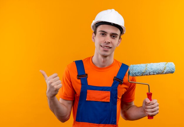 Homme jeune constructeur portant un uniforme de construction et un casque de sécurité faisant des pouces heureux et tenant une brosse à rouleau