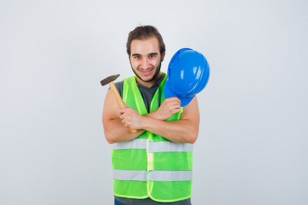 Photo gratuite homme jeune constructeur tenant un casque et un marteau tout en croisant les bras sur la poitrine en uniforme de vêtements de travail et à la joyeuse. vue de face.