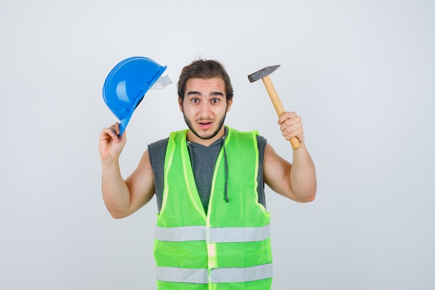 Homme Jeune Constructeur Tenant Un Casque Et Un Marteau En Uniforme De Vêtements De Travail Et à La Perplexité, Vue De Face.