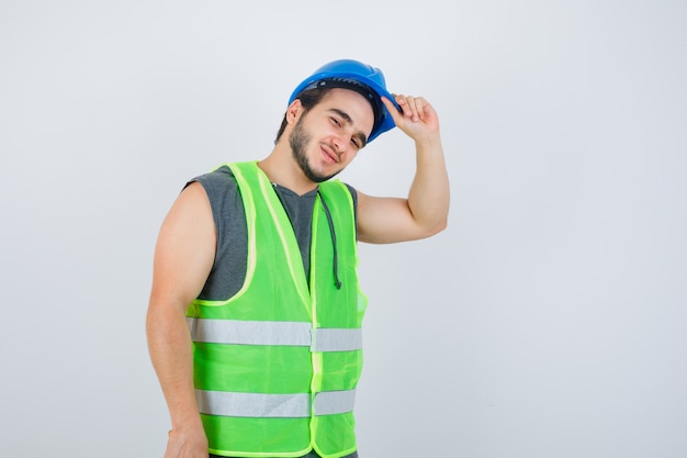 Homme jeune constructeur tenant la main sur le casque en uniforme de vêtements de travail et à la joyeuse. vue de face.