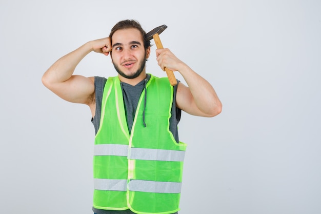 Homme jeune constructeur tenant le marteau et le poing sur la tête en uniforme de vêtements de travail et à la joyeuse vue de face.