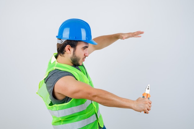 Homme jeune constructeur tenant des pinces tout en faisant semblant d'attraper quelque chose en uniforme de vêtements de travail et à la confiance en soi. vue de face.